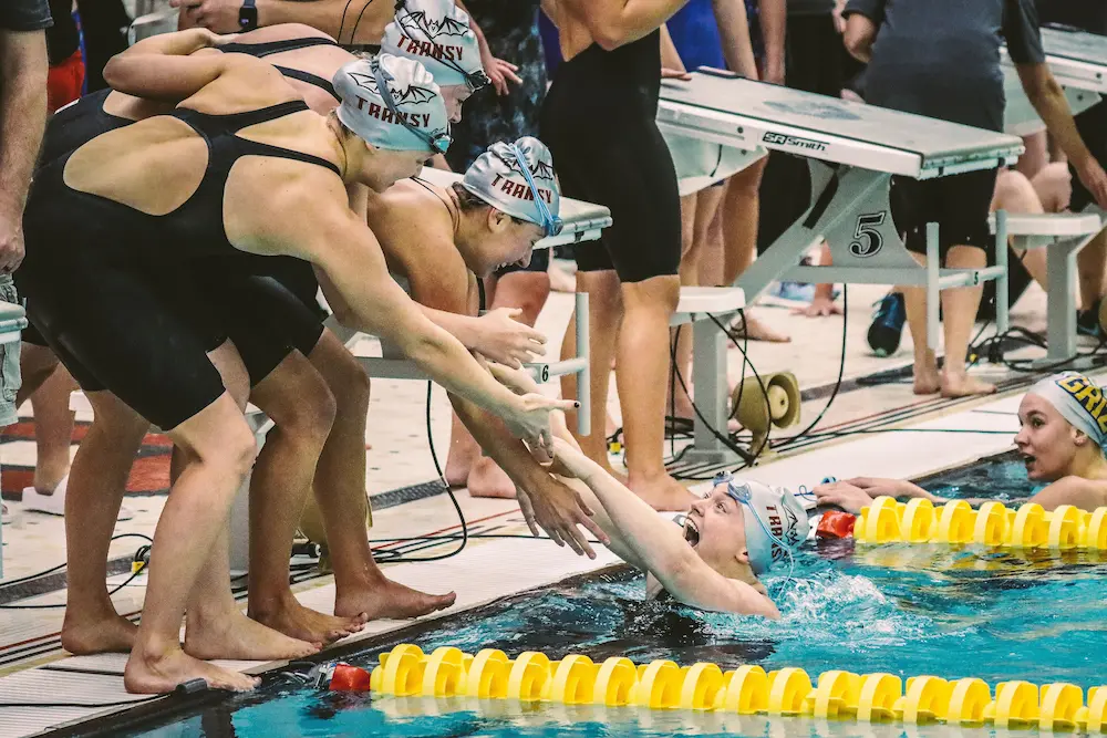 Team-mates reaching down to congratulate a swimmer at the wall during a busy swimming meet